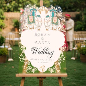 May include: A clear acrylic wedding sign on a wooden easel featuring two illustrated elephants with teal and gold decorative blankets. The text reads: ROHAN & AANYA Wedding FOREVER INLOVE 12.15.2024, surrounded by intricate gold floral filigree borders.