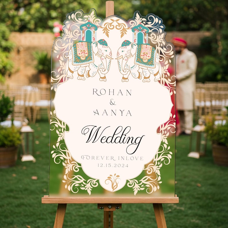 May include: A clear acrylic wedding sign on a wooden easel featuring two illustrated elephants with teal and gold decorative blankets. The text reads: ROHAN & AANYA Wedding FOREVER INLOVE 12.15.2024, surrounded by intricate gold floral filigree borders.