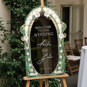 May include: An arched wedding sign with a black centre featuring the white text "WELCOME TO OUR WEDDING", "Amelia AND John", and "10.15.25". The frame is decorated with a green and cream stained glass pattern and white rose illustrations.