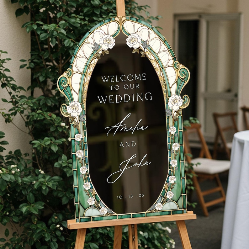 May include: An arched wedding sign with a black centre featuring the white text "WELCOME TO OUR WEDDING", "Amelia AND John", and "10.15.25". The frame is decorated with a green and cream stained glass pattern and white rose illustrations.