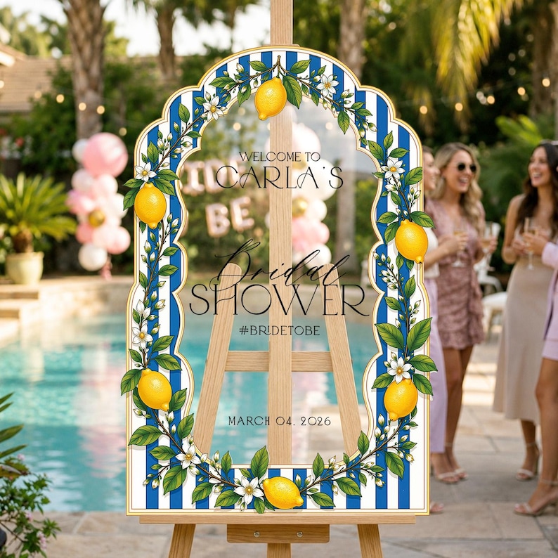 May include: A vertical arched sign on a wooden easel featuring a blue and white striped background with a border of yellow lemons and white blossoms. The text reads: WELCOME TO CARLA'S Bridal SHOWER #BRIDETOBE MARCH 04, 2026 in black lettering.