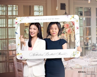 May include: Two women hold a white photo booth frame with floral accents. The frame says "We're so glad you're here!" and "Nathalie's Bridal Shower".