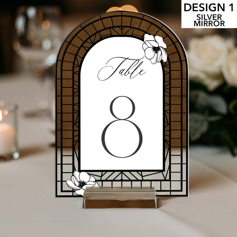 May include: Silver mirror table number with an arched top and a black geometric design. The number 8 is displayed in a large, bold font. The word "Table" is written in elegant script. Floral accents adorn the top and bottom.