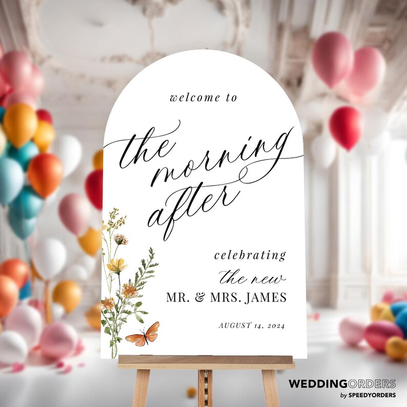 May include: A white wedding sign on a wooden easel, arched at the top, with the words "welcome to the morning after" in elegant script. It includes the names "MR. & MRS. JAMES" and the date "AUGUST 14, 2024". Floral and butterfly illustrations adorn the left side. Balloons in various colors are in the background.