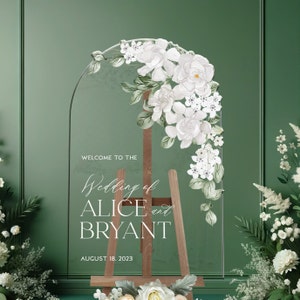 May include: A clear arched acrylic wedding sign featuring white floral illustrations and the text: WELCOME TO THE Wedding of ALICE and BRYANT AUGUST 18. 2023. The sign is displayed on a wooden easel, decorated with white flowers at the top right.