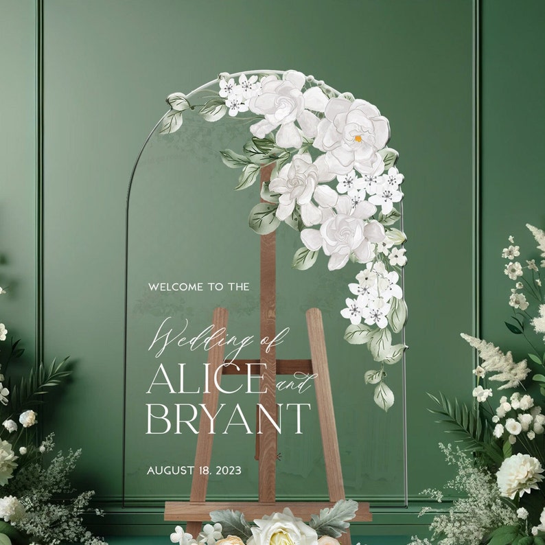 May include: A clear arched acrylic wedding sign featuring white floral illustrations and the text: WELCOME TO THE Wedding of ALICE and BRYANT AUGUST 18. 2023. The sign is displayed on a wooden easel, decorated with white flowers at the top right.