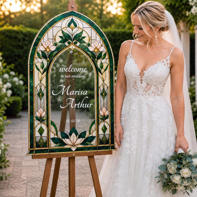 May include: An arched wedding welcome sign featuring a stained glass frame with green and cream floral patterns. The clear center panel displays the text: welcome to our wedding, Marisa & Arthur, 05-02-26. The sign is mounted on a wooden easel.