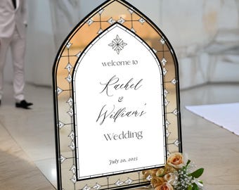 May include: An arched wedding welcome sign with a black frame and a stained glass-inspired border. The center features the text "welcome to Rachel & William's Wedding July 20, 2025" in black lettering on a white background, accented by a small floral motif.