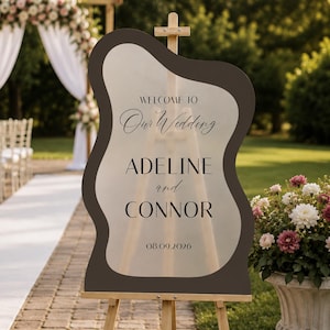 May include: A wavy-edged, dark brown and frosted acrylic wedding sign on a wooden easel. The text reads: WELCOME TO Our Wedding ADELINE and CONNOR 08.09.2026. The sign features a central translucent area with dark lettering and a solid dark border.