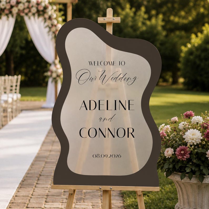 May include: A wavy-edged, dark brown and frosted acrylic wedding sign on a wooden easel. The text reads: WELCOME TO Our Wedding ADELINE and CONNOR 08.09.2026. The sign features a central translucent area with dark lettering and a solid dark border.