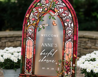 May include: An arched sign with a stained glass border in shades of red, pink, and green, featuring white floral branches and butterflies. The center text reads: WELCOME TO ANNE'S bridal shower 10.15.25. The sign has a frosted, semi-transparent finish.