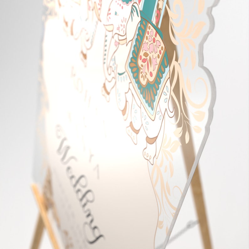 May include: A clear acrylic wedding sign featuring a printed illustration of an elephant adorned with teal, pink, and gold decorative patterns. The sign includes elegant script text that reads Wedding, displayed on a light-colored wooden easel stand.