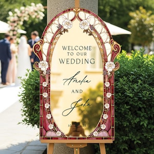 May include: A decorative wedding sign with a stained-glass design. The sign reads "Welcome to our Wedding" above the names "Amelia and John" and the date "10.25". The sign is framed with burgundy and gold accents and white roses.