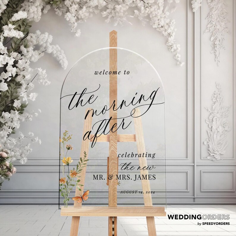 May include: Clear acrylic wedding sign on a wooden easel. The sign reads "welcome to the morning after" in elegant script, with the names "MR. & MRS. JAMES" and the date "AUGUST 14, 2024". Floral and butterfly accents add a decorative touch.