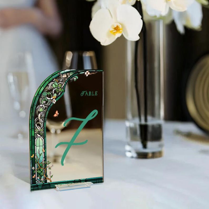 May include: A green and gold mirrored table number with a floral design and the word "TABLE" printed on it. The number "7" is written in green on the mirror.