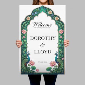 Op de afbeelding: Een rechthoekig welkomstbord voor een bruiloft met een boogvormige rand versierd met groene bladeren, blauwe en cr&egrave;mekleurige bloemen en roze lotusbloemen. Twee pauwen met blauwe halzen en groene staarten flankeren de tekst: Welcome to the wedding of Dorothy & Lloyd, June 6, 2026.
