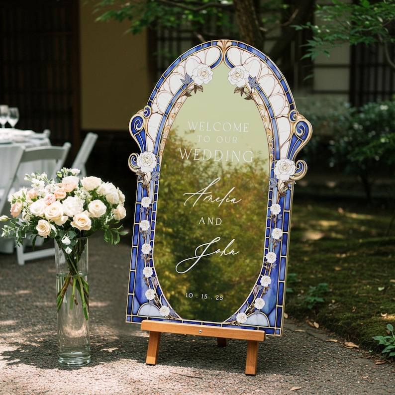 May include: A decorative wedding sign with a stained glass design. The sign reads "Welcome to our Wedding" with the names "Amelia and John" and the date "10.15.25". The sign is supported by a wooden easel and features white roses.