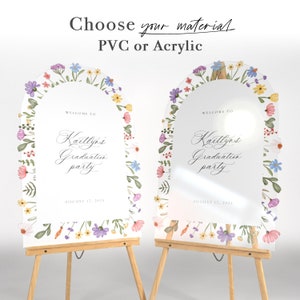 May include: Two arched signs for a graduation party, one opaque and one transparent, featuring a floral border with purple, blue, yellow, and pink flowers. The text reads "Choose your material PVC or Acrylic" and "WELCOME TO Kaitlyn's Graduation party AUGUST 17, 2024".