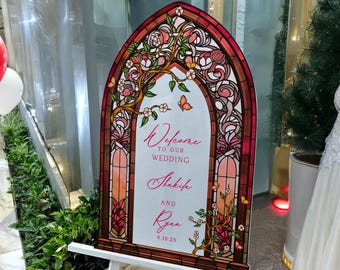 May include: An arched wedding welcome sign featuring a stained glass window design with red, pink, and orange tones. The sign displays the text Welcome to our wedding, Shakifa and Ryan, 6.15.25, surrounded by illustrated floral branches and butterflies.