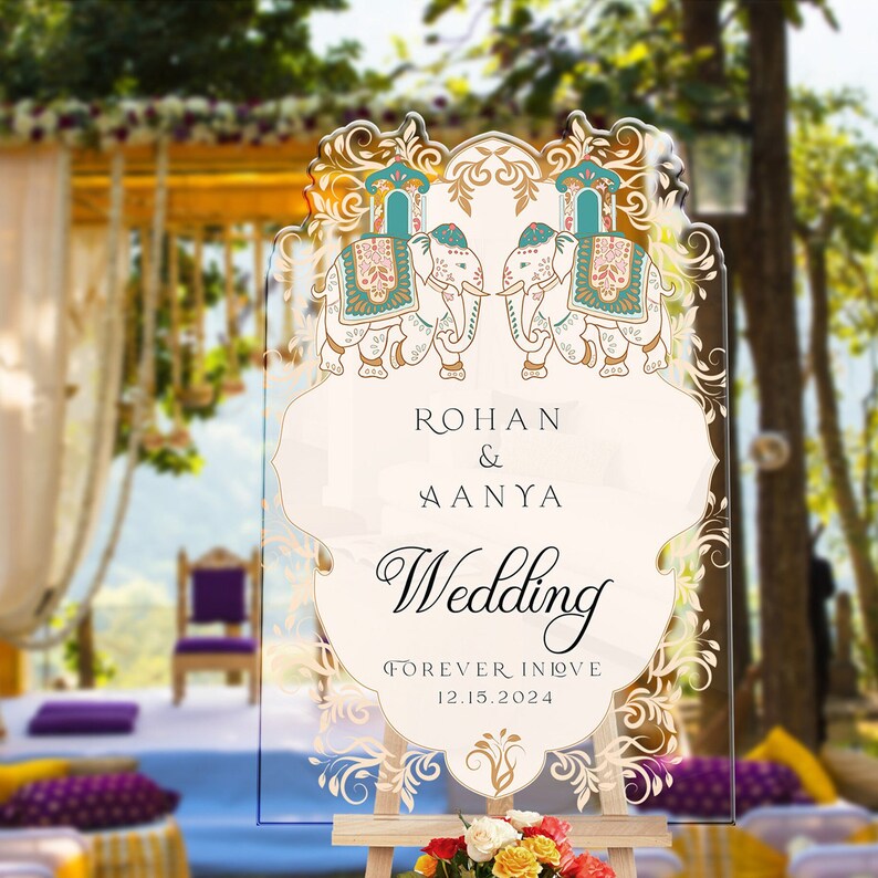 May include: A clear acrylic wedding sign with an arched top, featuring two illustrated elephants in teal, pink, and gold. The text reads: ROHAN & AANYA, Wedding, FOREVER IN LOVE, 12.15.2024. The sign is decorated with intricate gold filigree borders.
