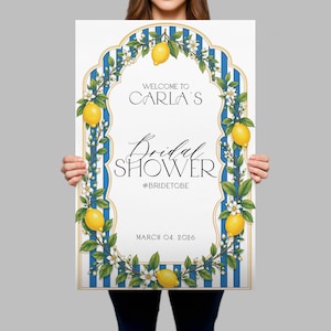 May include: A rectangular sign featuring a white center with blue vertical stripes on the border. The design includes yellow lemons and white flowers. Text reads: WELCOME TO CARLA'S Bridal SHOWER #BRIDETOBE MARCH 04, 2026. The sign has a gold-toned frame.