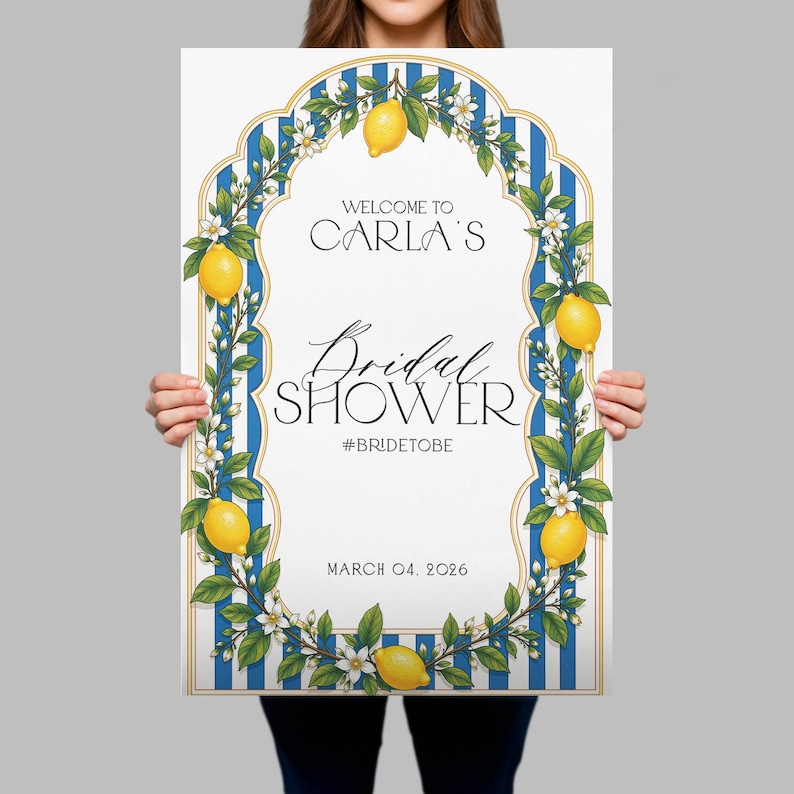 May include: A rectangular sign featuring a white center with blue vertical stripes on the border. The design includes yellow lemons and white flowers. Text reads: WELCOME TO CARLA'S Bridal SHOWER #BRIDETOBE MARCH 04, 2026. The sign has a gold-toned frame.