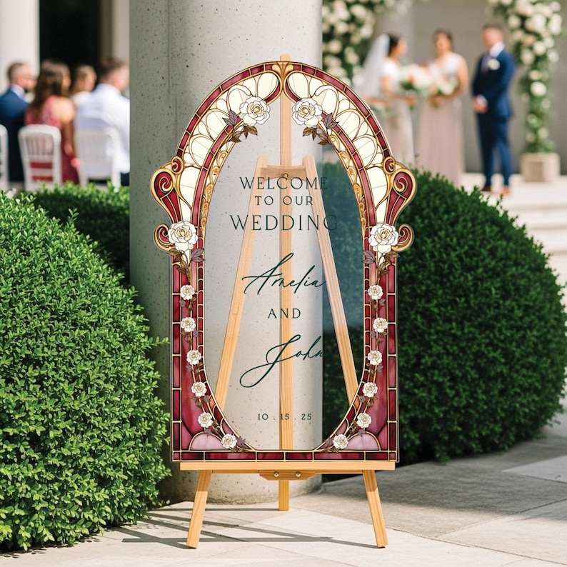 May include: A decorative wedding sign with a stained-glass design. The sign features an oval frame with floral accents and the words "Welcome to our Wedding" along with the names Amelia and John and the date 10.15.25. The sign is displayed on a wooden easel.