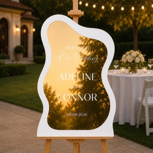 May include: A wavy-edged gold mirror wedding sign with a white frame, mounted on a wooden easel. The text reads: WELCOME TO Our Wedding ADELINE and CONNOR 08.09.2026. The reflective surface shows a sunset sky and tree silhouettes.