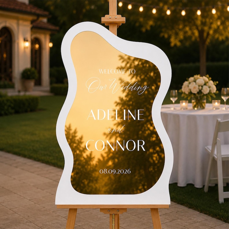 May include: A wavy-edged gold mirror wedding sign with a white frame, mounted on a wooden easel. The text reads: WELCOME TO Our Wedding ADELINE and CONNOR 08.09.2026. The reflective surface shows a sunset sky and tree silhouettes.