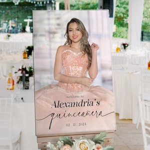 May include: A pink and white welcome sign for a quincea&ntilde;era. The sign features a photo of a young woman in a pink dress and the text "Welcome to Alexandria's Quincea&ntilde;era 02.11.2024".