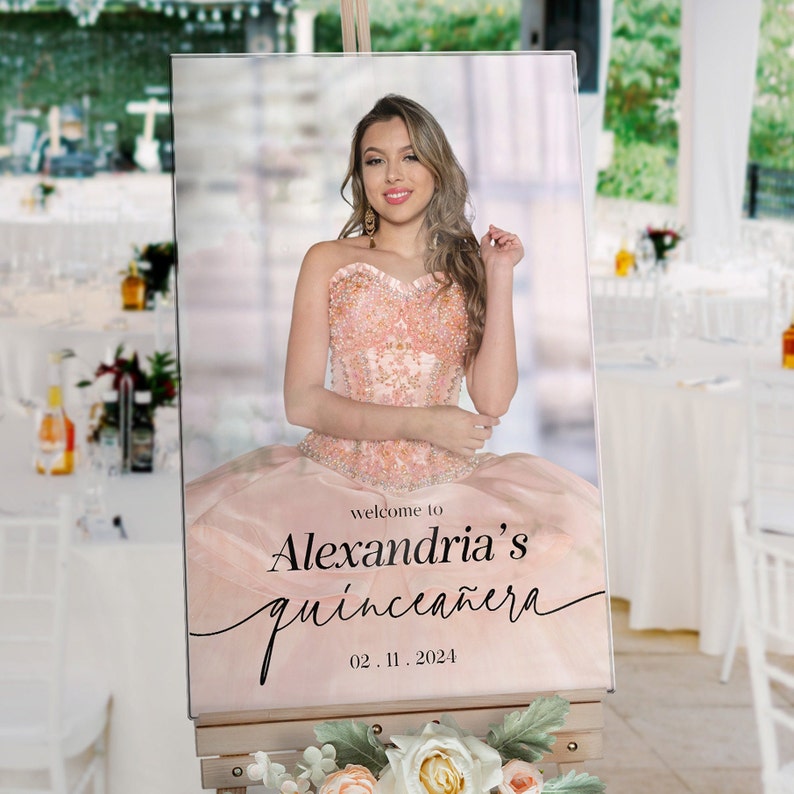 May include: A pink and white welcome sign for a quincea&ntilde;era. The sign features a photo of a young woman in a pink dress and the text "Welcome to Alexandria's Quincea&ntilde;era 02.11.2024".