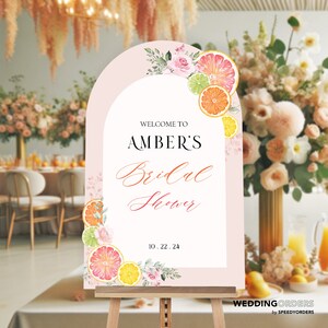 May include: A pink and white welcome sign with a floral design and citrus fruit slices. The sign reads "Welcome to Amber's Bridal Shower 10.22.24".