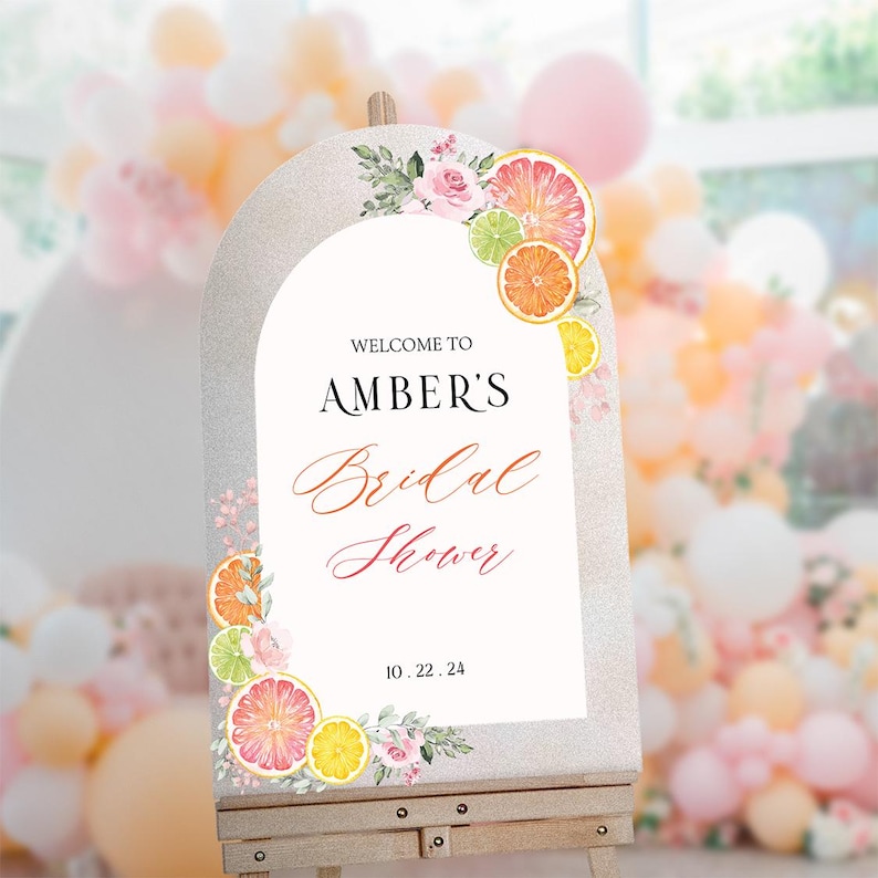 May include: A white welcome sign with a silver glitter background and a floral design featuring pink roses and citrus fruit slices. The sign reads "Welcome to Amber's Bridal Shower 10.22.24".