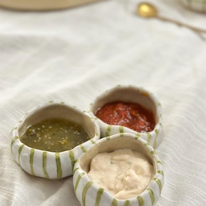 May include: A set of three small, rounded ceramic bowls with an irregular, hand-formed shape. Each bowl features a white base with vertical, hand-painted light green stripes. The bowls are filled with green salsa, red salsa, and a creamy white dip.