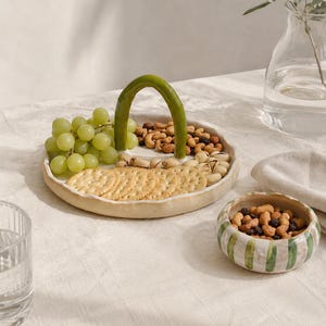 May include: A round, off-white ceramic serving tray with a high, arched green handle, divided into sections containing green grapes, crackers, and mixed nuts. Beside it sits a small, round ceramic bowl with vertical green stripes.