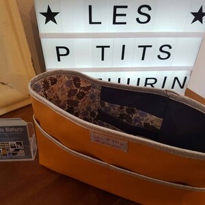 Pode incluir: Uma sacola laranja com acabamento cinza e interior estampado floral. A sacola tem dois bolsos externos e uma etiqueta que diz "Les P'tits Mathurin". Uma placa ao fundo exibe o texto "LES PTITS MATHURIN".