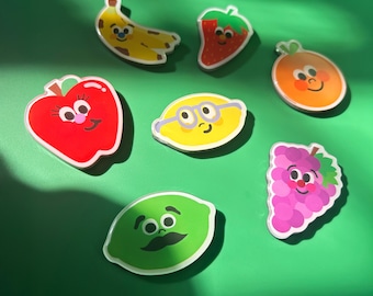 May include: A collection of colorful fruit-shaped stickers with smiling faces. The stickers include a yellow banana, red apple, green lime, orange, strawberry, lemon, and purple grapes, all with white outlines, set against a green background.