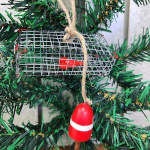 May include: A Christmas bauble featuring a red and white buoy and a wire lobster pot. The bauble is hanging from a brown twine string on a green artificial Christmas tree.