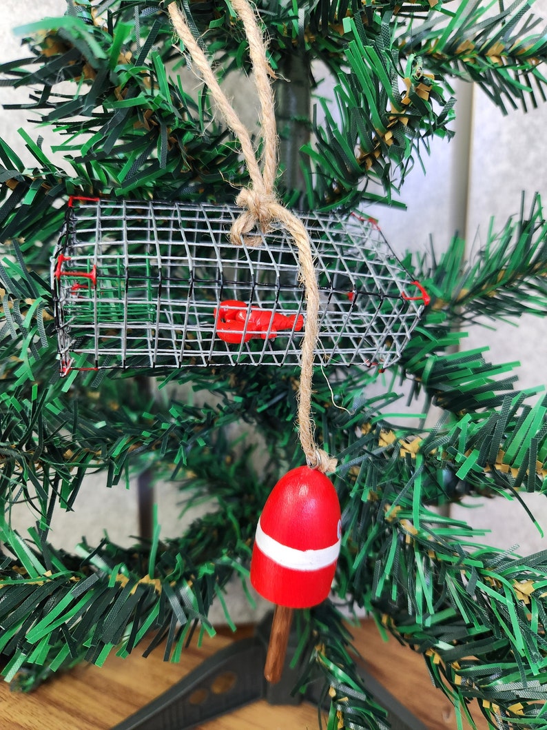 May include: A Christmas bauble featuring a red and white buoy and a wire lobster pot. The bauble is hanging from a brown twine string on a green artificial Christmas tree.