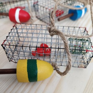 May include: A set of three miniature lobster trap ornaments featuring a red lobster, a red and white buoy, and a yellow and green buoy. The traps are made of wire mesh and have a rustic look.