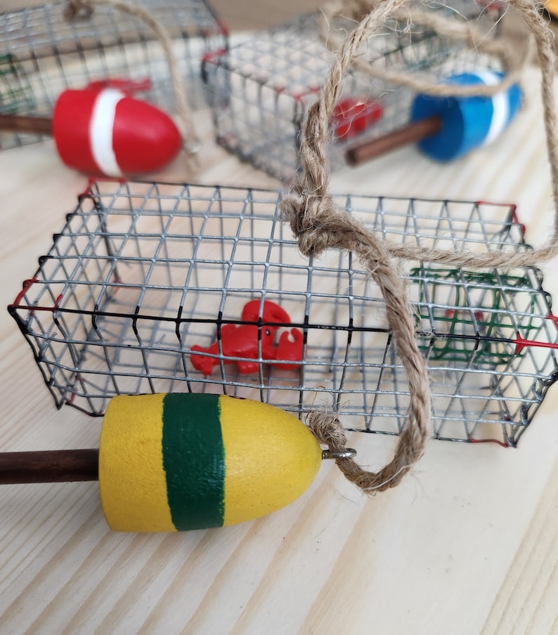 May include: A set of three miniature lobster trap ornaments featuring a red lobster, a red and white buoy, and a yellow and green buoy. The traps are made of wire mesh and have a rustic look.