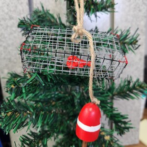 May include: A red and white buoy ornament with a wire lobster trap hanging from it. The ornament is made of wood and is decorated with a rope.
