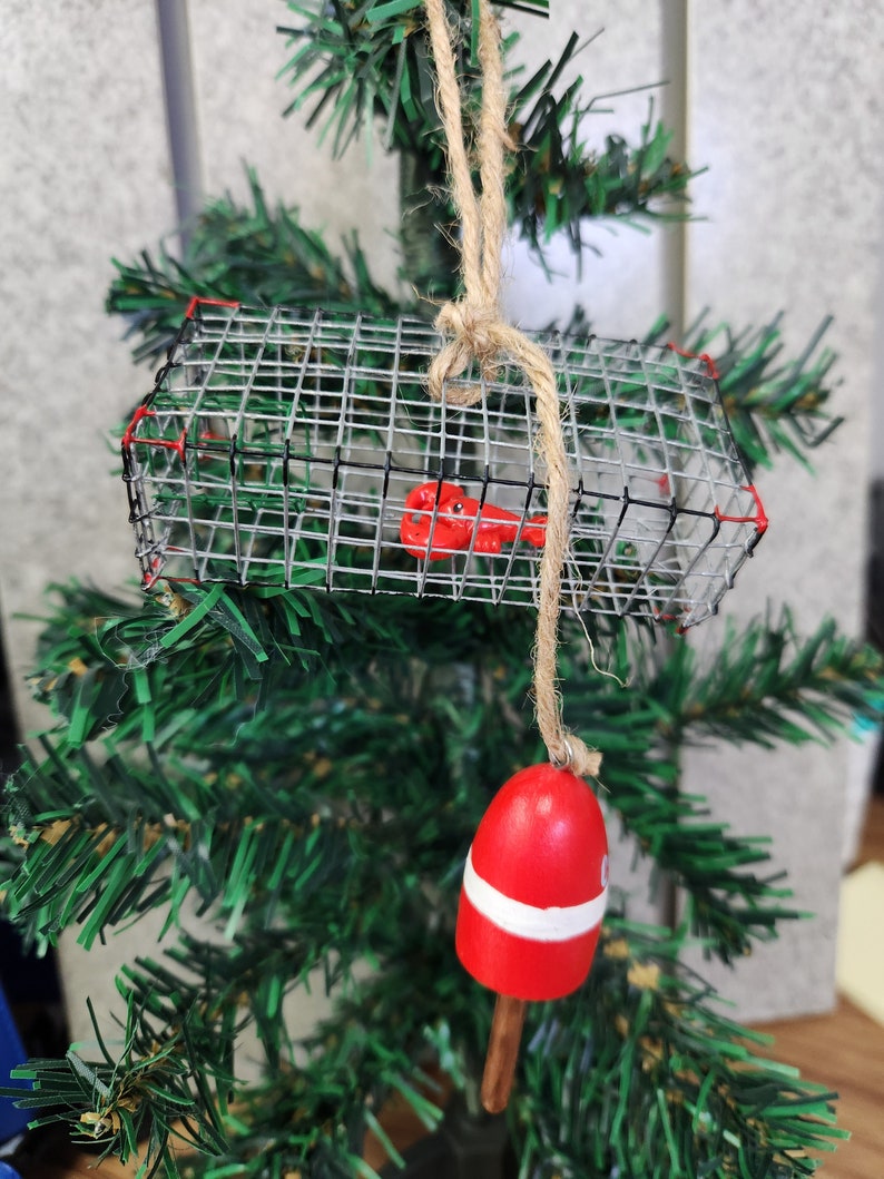 May include: A red and white buoy ornament with a wire lobster trap hanging from it. The ornament is made of wood and is decorated with a rope.