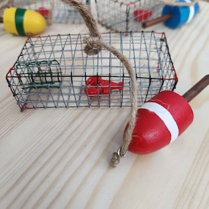 May include: A set of four miniature lobster trap ornaments featuring a red lobster, a red and white buoy, and a green and yellow buoy. The traps are made of wire mesh and are hung with twine.