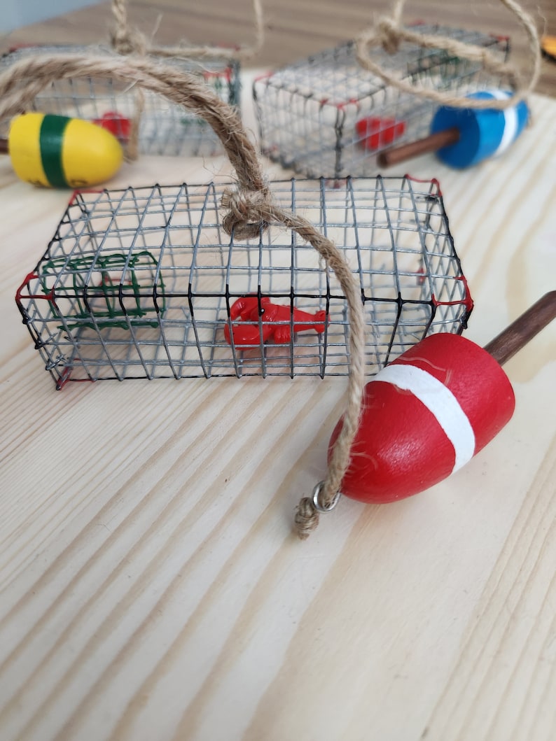 May include: A set of four miniature lobster trap ornaments featuring a red lobster, a red and white buoy, and a green and yellow buoy. The traps are made of wire mesh and are hung with twine.