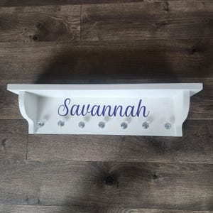 May include: White wooden shelf with the name "Savannah" written in purple lettering. The shelf has seven crystal knobs for hanging items.