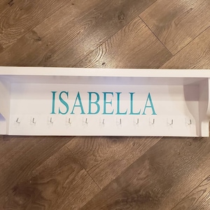 May include: White wooden shelf with a teal inscription that reads "ISABELLA" and multiple hooks.