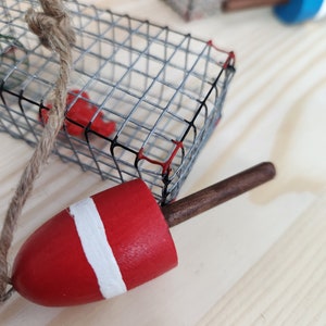 May include: A red and white buoy with a brown wooden handle attached to a wire cage with a rope. The cage is made of metal and has a red and black design.