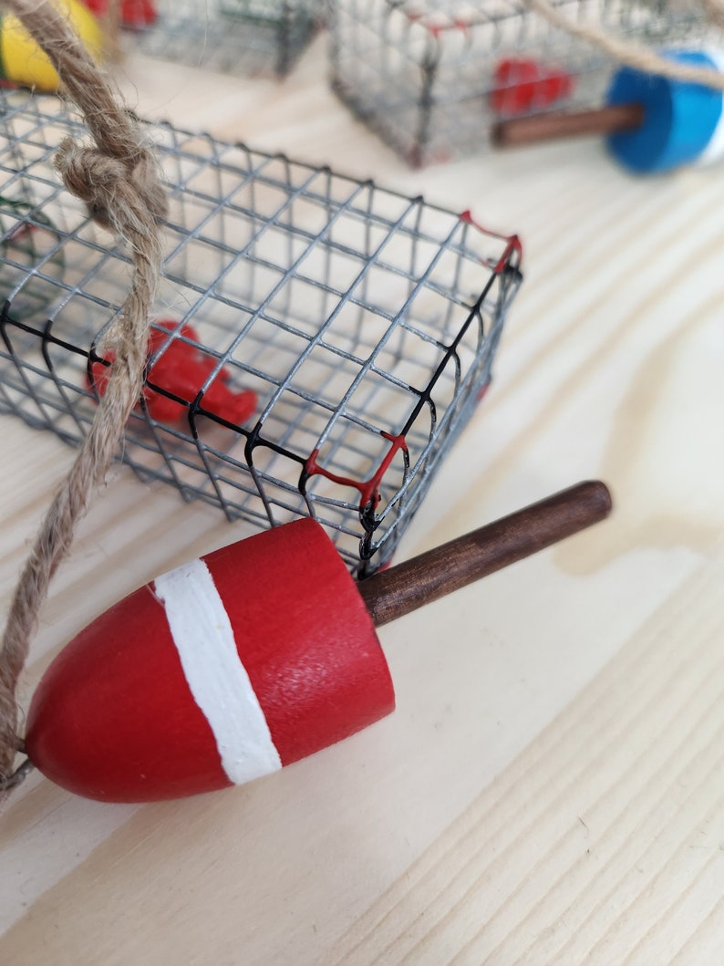 May include: A red and white buoy with a brown wooden handle attached to a wire cage with a rope. The cage is made of metal and has a red and black design.