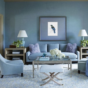 May include: A light blue living room with a light blue sofa, two chairs, a coffee table, and a rug. The walls are painted a light blue color. There is a framed print of a bird on the wall. The coffee table has a glass top and a white ottoman.